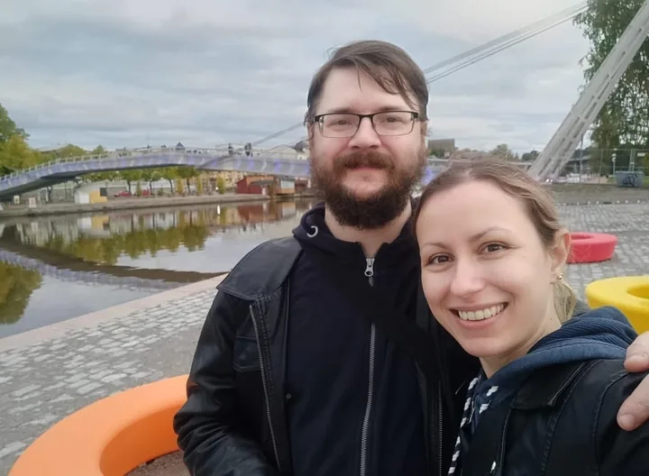 Angelina and Alain smiling into the camera by a river, with a footbridge behind them.